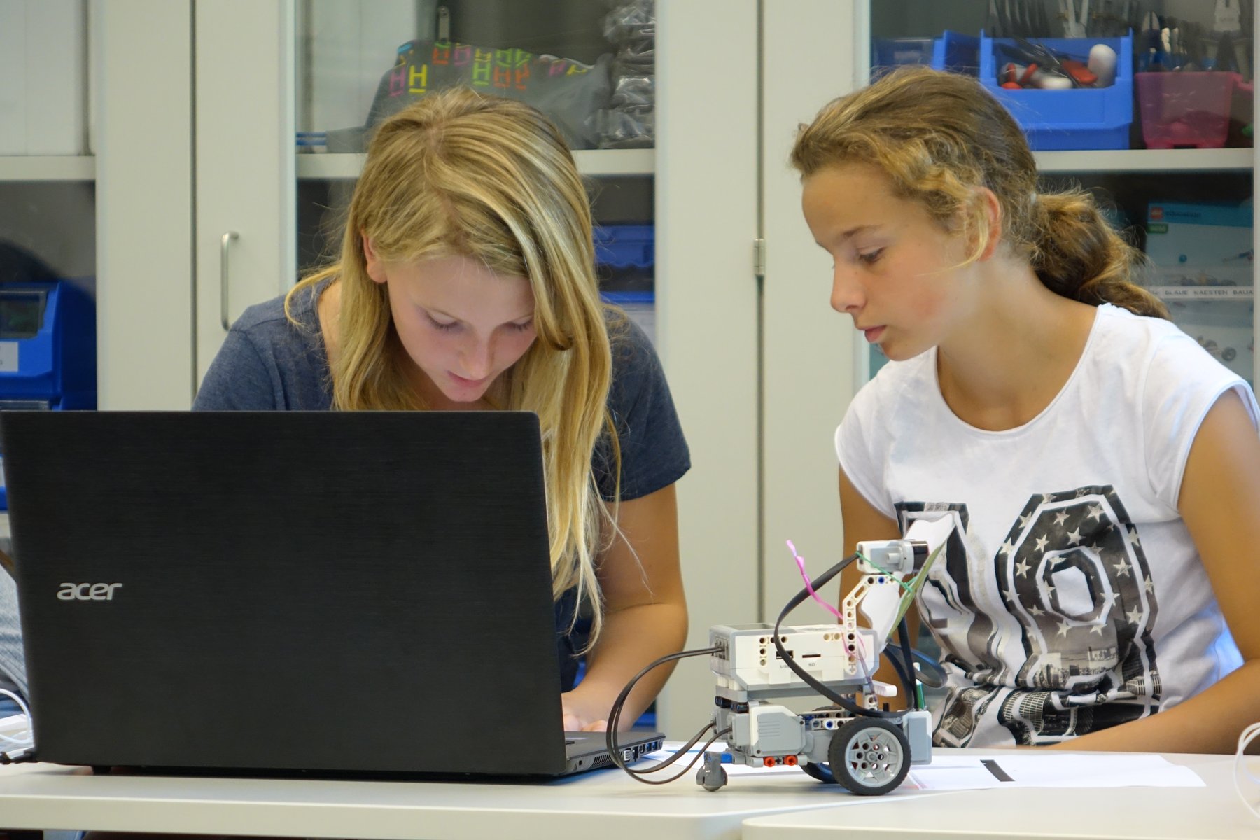 Zwei junge Frauen am Tisch vor Labtop mit kleinem Roboter auf Rädern vor sich