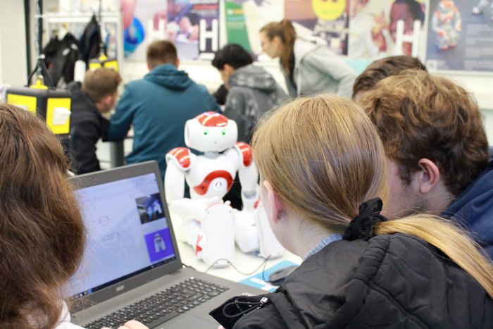 Blick von hinten auf Schülerinnen und Schüler beim Programmieren. Ihr Blick fällt auf ein Notebook, hinter dem ein NAO-Roboter sitzt. Der NAO-Roboter ist den Schülerinnen und Schülern zugewandt.