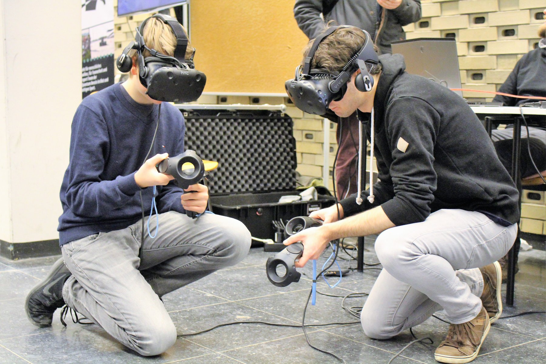 Zwei junge Männer mit VR Brillen knien am Boden und probieren Technik aus 